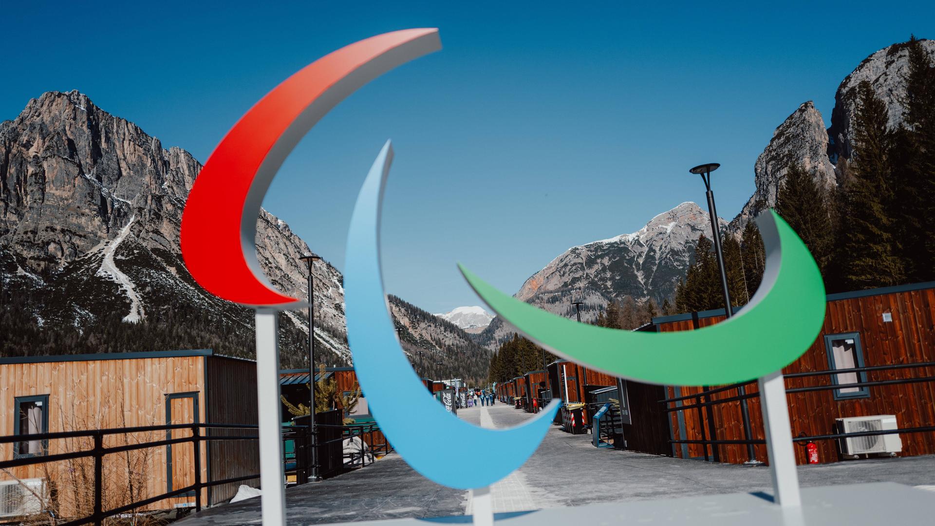 Das Logo der Paralympischen Spiele steht vor einer Bergkulisse. Es besteht aus drei geschwungenen Formen, die wie Pinselstriche oder Sicheln aussehen. Die Farben sind Rot, Blau und Grün.