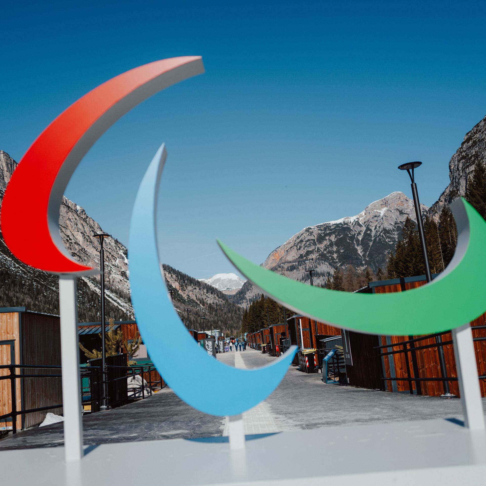 Das Logo der Paralympischen Spiele steht vor einer Bergkulisse. Es besteht aus drei geschwungenen Formen, die wie Pinselstriche oder Sicheln aussehen. Die Farben sind Rot, Blau und Grün.