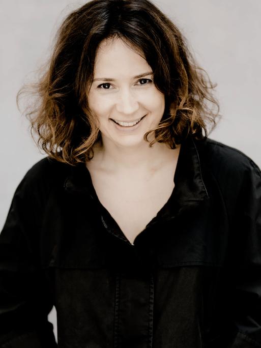 Eine Frau mit kurzen dunklen Locken blickt lächelnd in die Kamera. Sie trägt ein schwarzes Oberteuil vor weißem Hintergrund