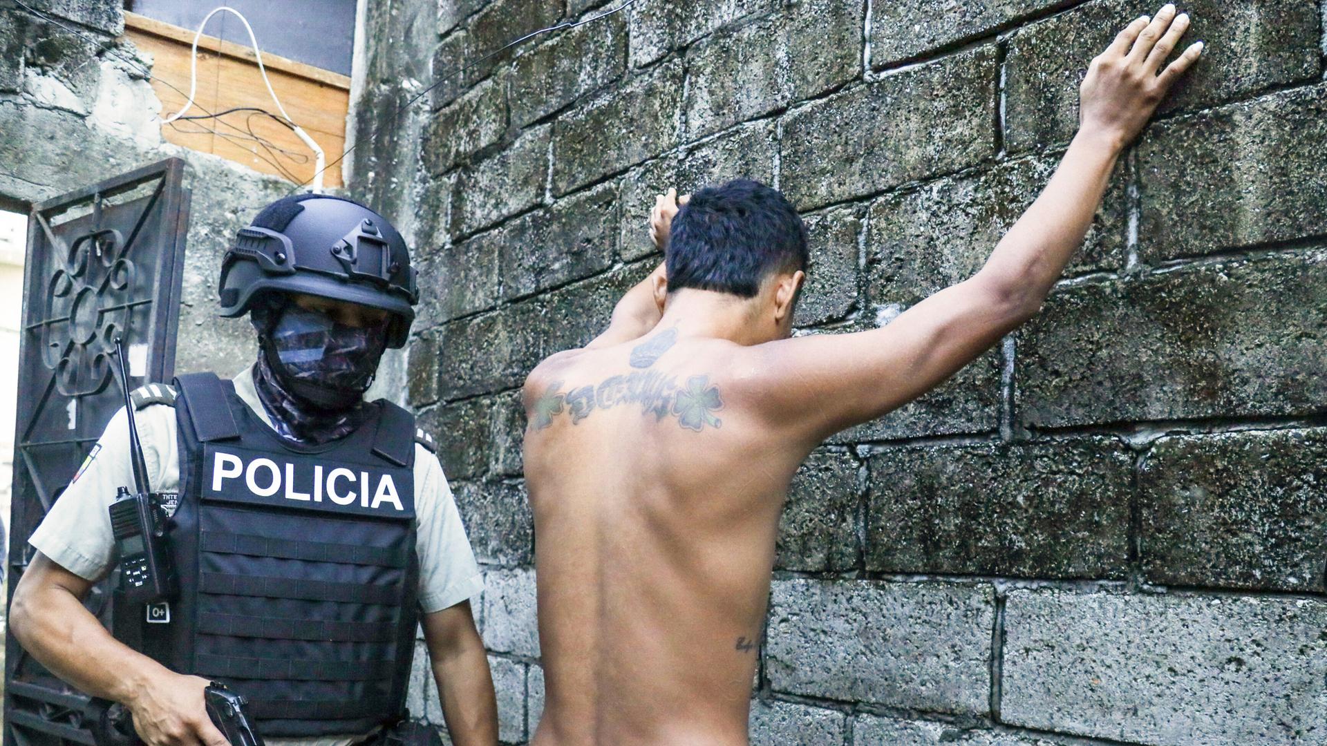 Ein Bewohner steht mit gespreizten Armen an einer Wand, während die Polizei nach Waffen oder Drogen sucht.