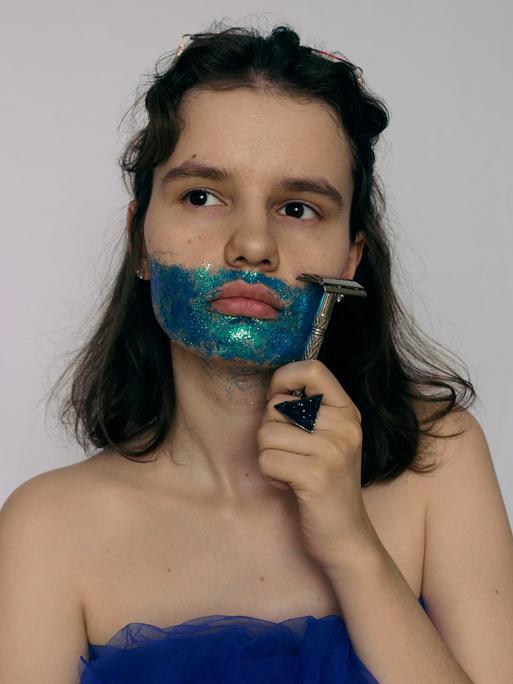 Eine Frau hält sich einen Rasierer an die Backe. Sie trägt einen Bart aus blauer, glitzernder Farbe und blickt zum Bildrand hin. Eine Frau hält sich einen Rasierer an die Backe. Sie trägt einen Bart aus blauer, glitzernder Farbe und blickt zum Bildrand hin.