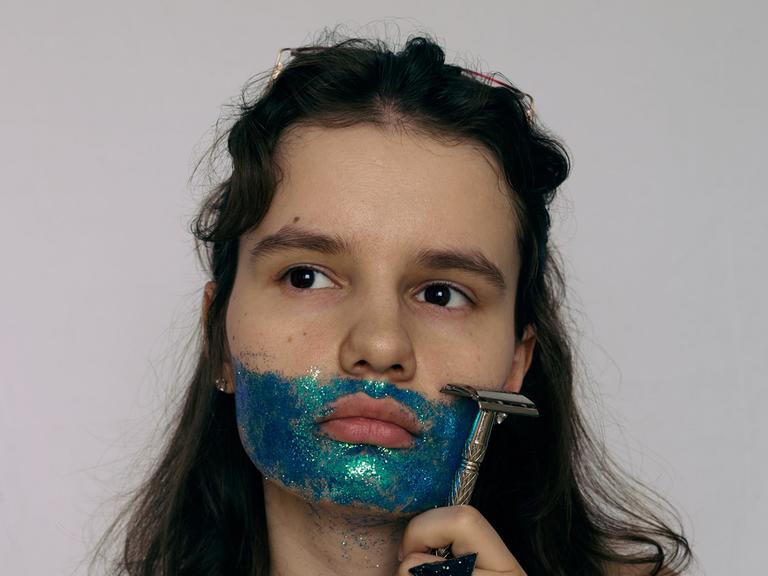 Eine Frau hält sich einen Rasierer an die Backe. Sie trägt einen Bart aus blauer, glitzernder Farbe und blickt zum Bildrand hin. Eine Frau hält sich einen Rasierer an die Backe. Sie trägt einen Bart aus blauer, glitzernder Farbe und blickt zum Bildrand hin.
