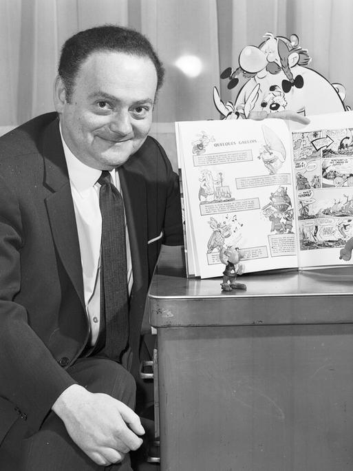 Schwarzweiß-Aufnahme von René Goscinny, der stolz einen Band seiner "Asterix"-Reihe präsentiert