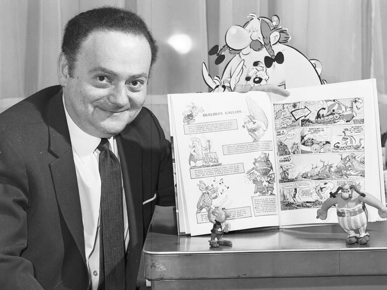 Schwarzweiß-Aufnahme von René Goscinny, der stolz einen Band seiner "Asterix"-Reihe präsentiert