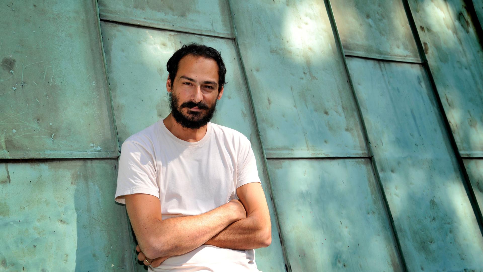 Mazen Kerbaj steht im Jahr 2015 vor einer türkisgrünen Blechkonstruktion. Der Künstler hat schwarze Haare und einen Bart, er trägt ein weißes T-Shirt.