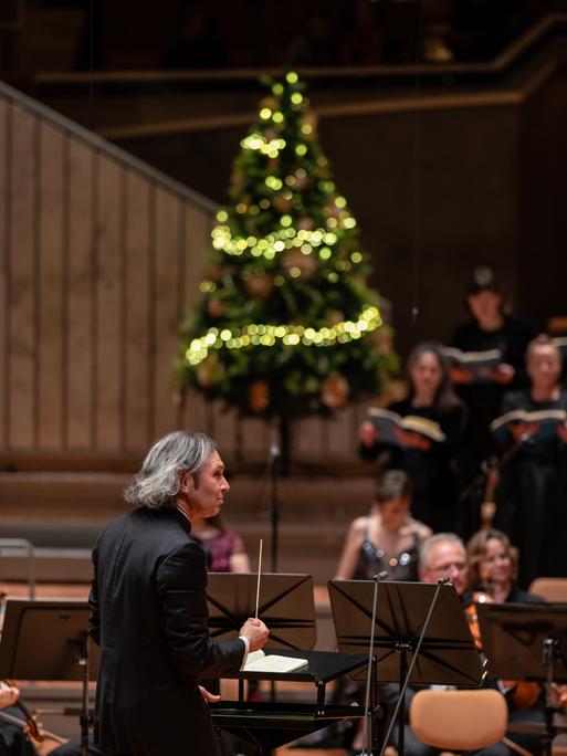 Vladimir Jurowski dirigiert Musiker und Sängerinnen, während auf der Bühne der Philharmonie Berlin ein Weihnachtsbaum steht.