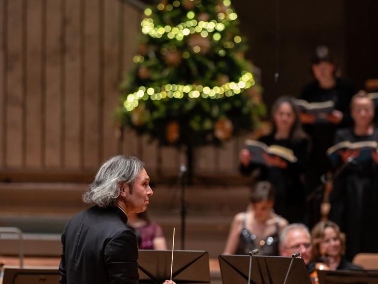 Vladimir Jurowski dirigiert Musiker und Sängerinnen, während auf der Bühne der Philharmonie Berlin ein Weihnachtsbaum steht.