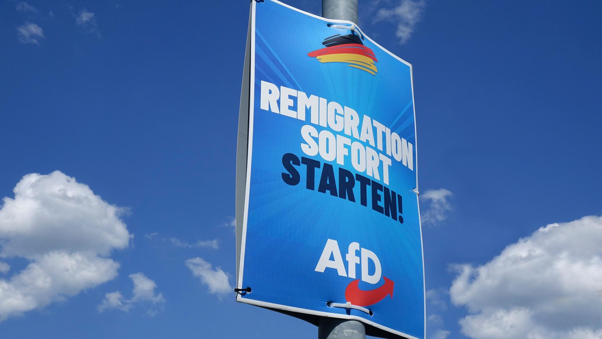 Das Plakat hängt an einem Pfosten unter einem blauen Himmel mit Wolken. Darauf steht: "Remigration sofort starten - AfD."