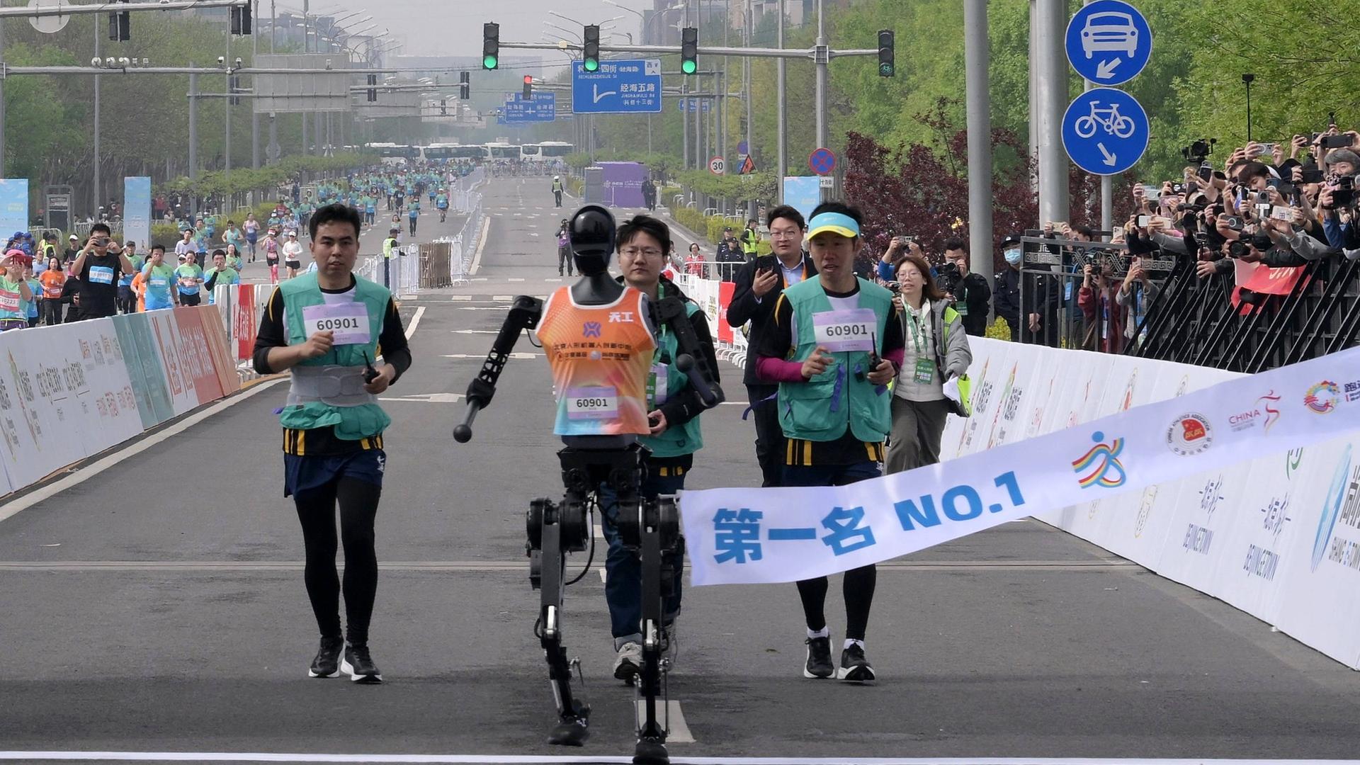 Ein humanoider Roboter läuft beim Halbmarathon in Peking durch das Ziel - hinter ihm sind menschliche Läufer zu sehen. Ein humanoider Roboter läuft beim Halbmarathon in Peking durch das Ziel - hinter ihm sind menschliche Läufer zu sehen.