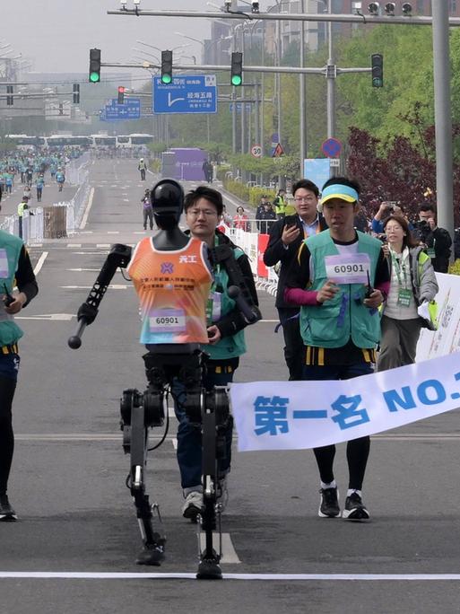 Ein humanoider Roboter läuft beim Halbmarathon in Peking durch das Ziel - hinter ihm sind menschliche Läufer zu sehen.