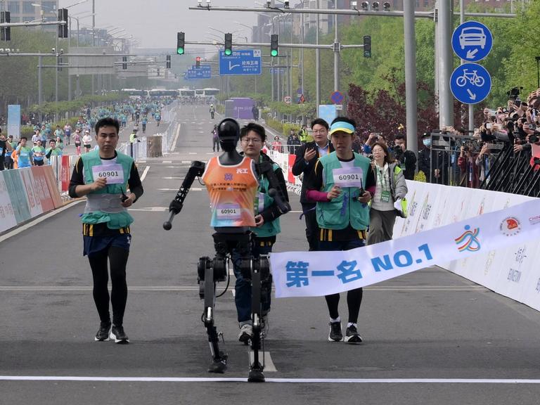 Ein humanoider Roboter läuft beim Halbmarathon in Peking durch das Ziel - hinter ihm sind menschliche Läufer zu sehen.