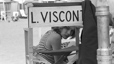 Schwarzweißfoto von Björn Andrésen am Set von "Der Tod in Venedig", der auf einem Stuhl mit der Aufschrift "L. Visconti" sitzt und durch die Lehne in die Kamera schaut.