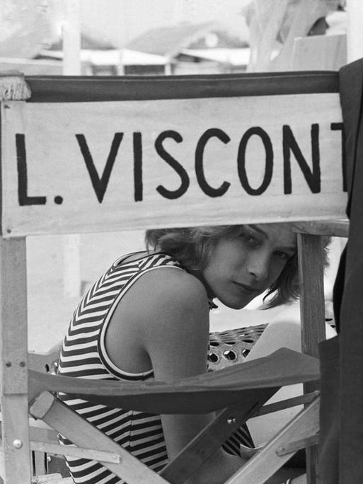 Schwarzweißfoto von Björn Andrésen am Set von "Der Tod in Venedig", der auf einem Stuhl mit der Aufschrift "L. Visconti" sitzt und durch die Lehne in die Kamera schaut.