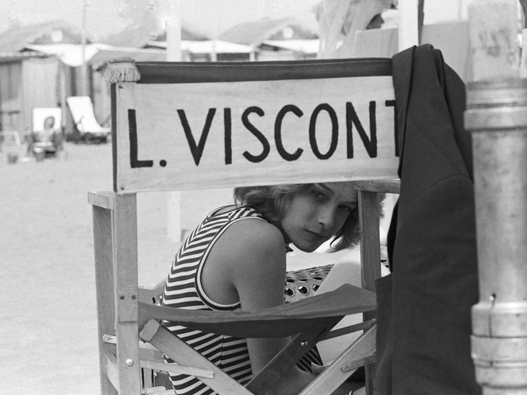 Schwarzweißfoto von Björn Andrésen am Set von "Der Tod in Venedig", der auf einem Stuhl mit der Aufschrift "L. Visconti" sitzt und durch die Lehne in die Kamera schaut.