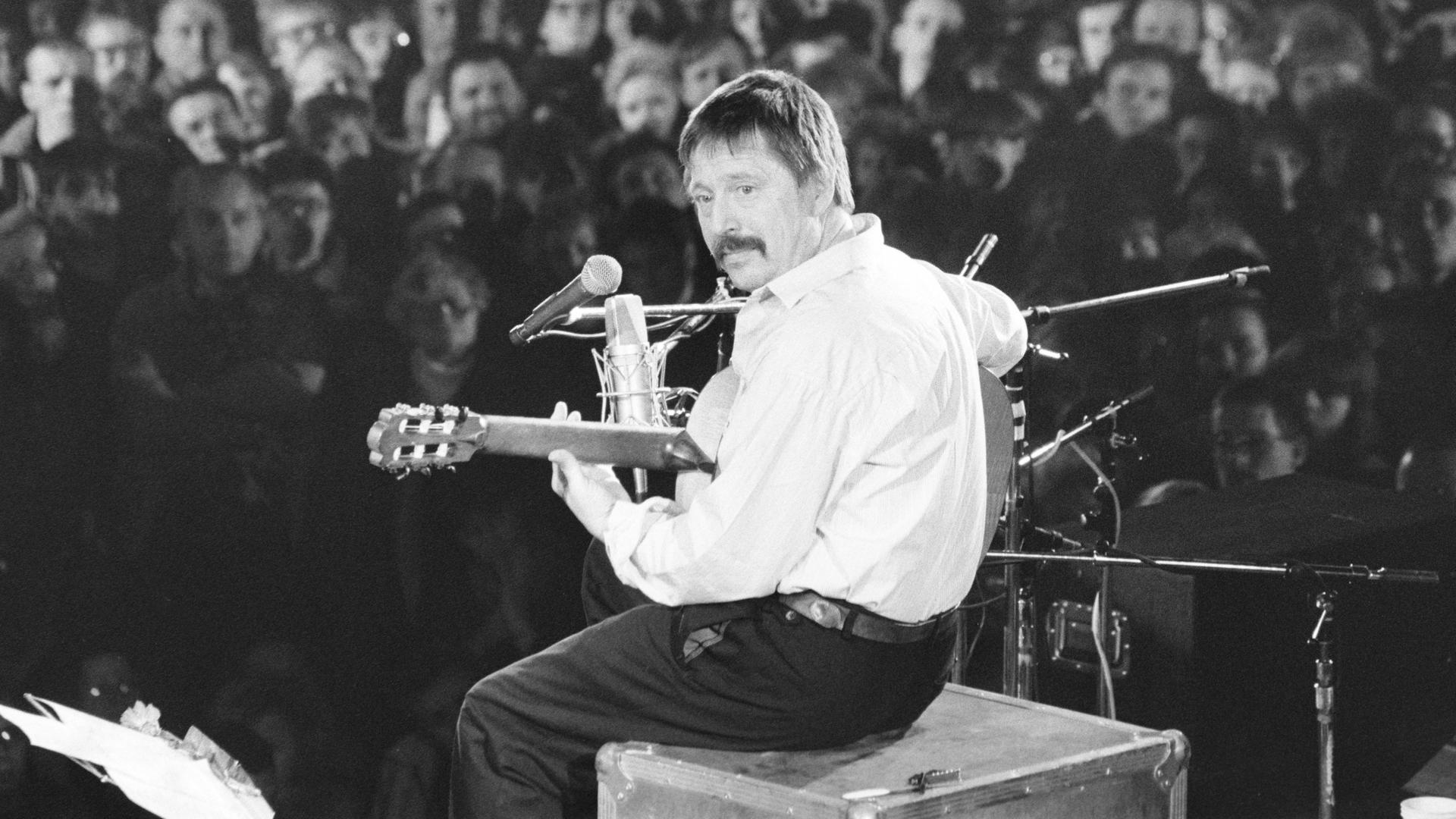 Das Schwarzweißfoto zeigt den Liedermacher Wolf Biermann, aufgenommen seitlich von hinten. Biermann sitzt auf einer Kiste auf der Bühne der Leipziger Messehalle, spielt Gitarre und singt in ein Mikrofon. Im Hintergrund sind zahlreiche Zuschauer zu sehen.