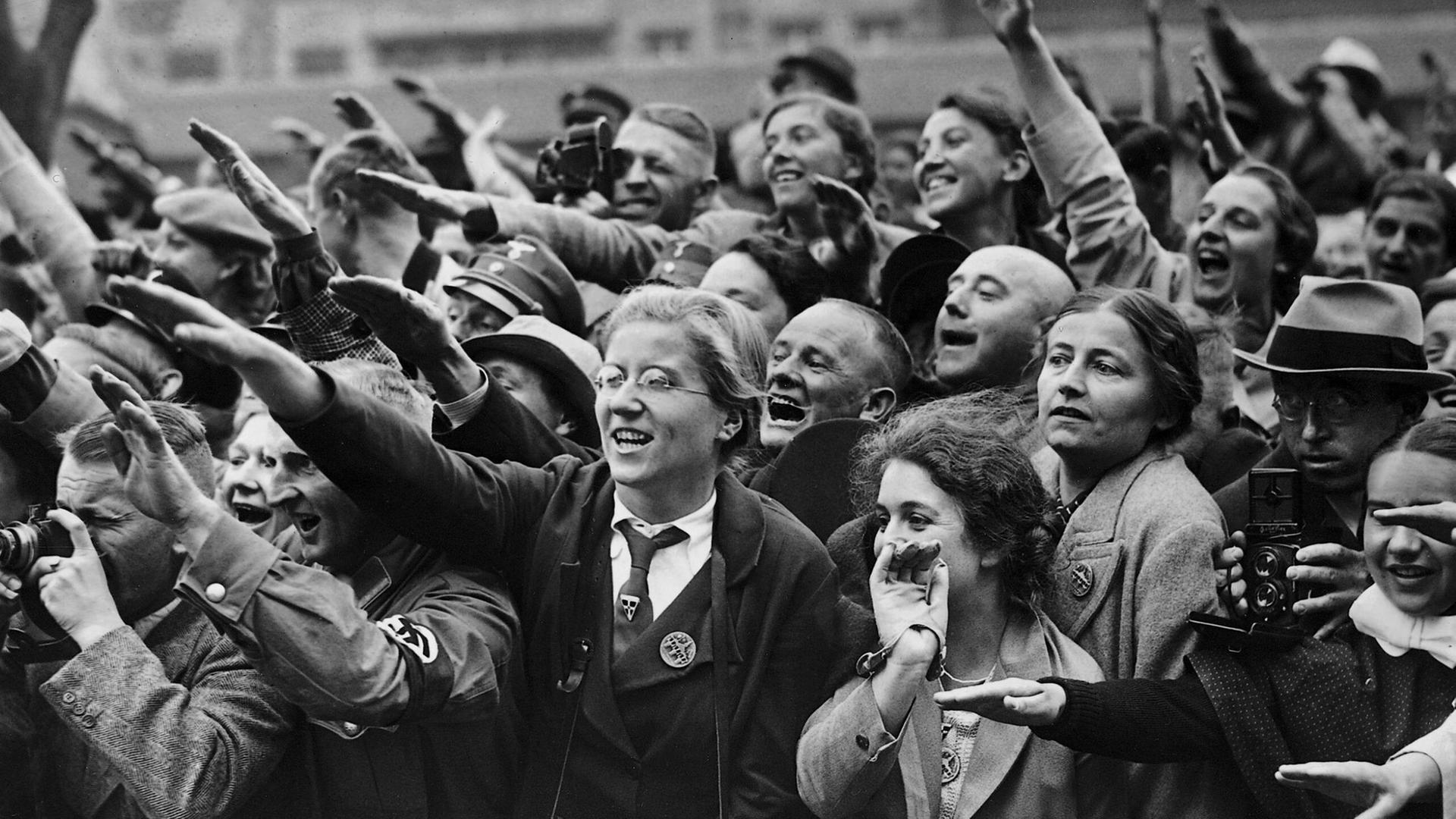 Schwarz-Weiß-Bild von begeisterten Frauen und Männern, die den Hitlergruß zeigen und  dicht gedrängt am Straßenrand stehen