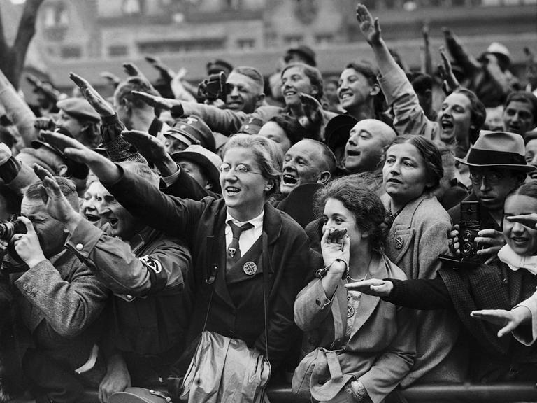 Schwarz-Weiß-Bild von begeisterten Frauen und Männern, die den Hitlergruß zeigen und  dicht gedrängt am Straßenrand stehen