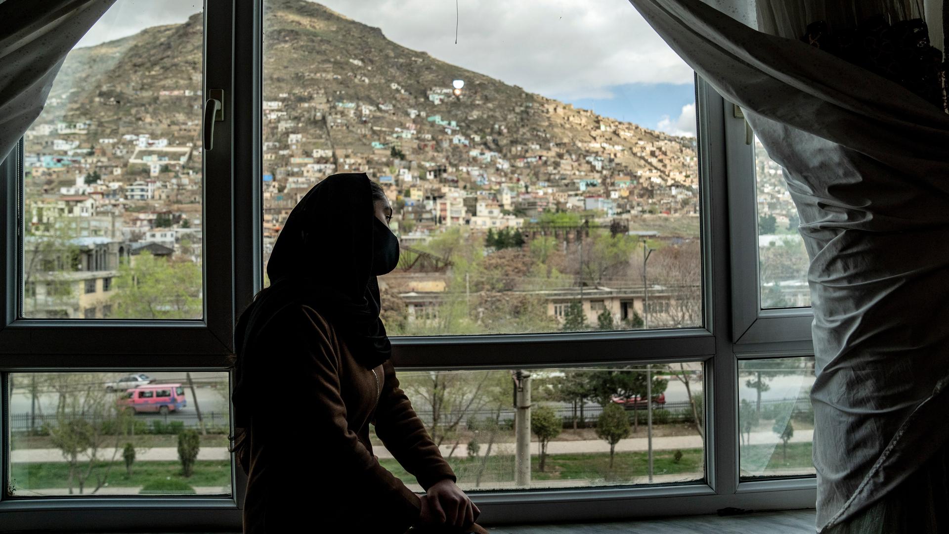 Schattenumriss einer jungen Frau in Afghanistan, die auf einer Fensterbank sitzt und hinausschaut. Im Hintergrund ist eine Stadt zu sehen. Schattenumriss einer jungen Frau in Afghanistan, die auf einer Fensterbank sitzt und hinausschaut. Im Hintergrund ist eine Stadt zu sehen.
