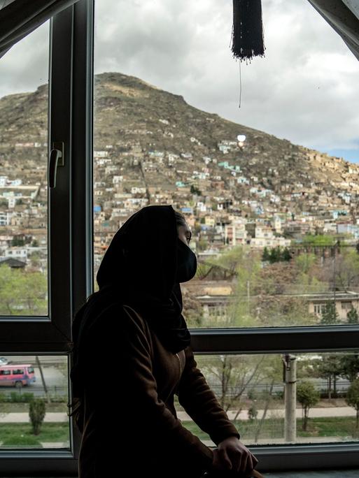 Schattenumriss einer jungen Frau in Afghanistan, die auf einer Fensterbank sitzt und hinausschaut. Im Hintergrund ist eine Stadt zu sehen. 