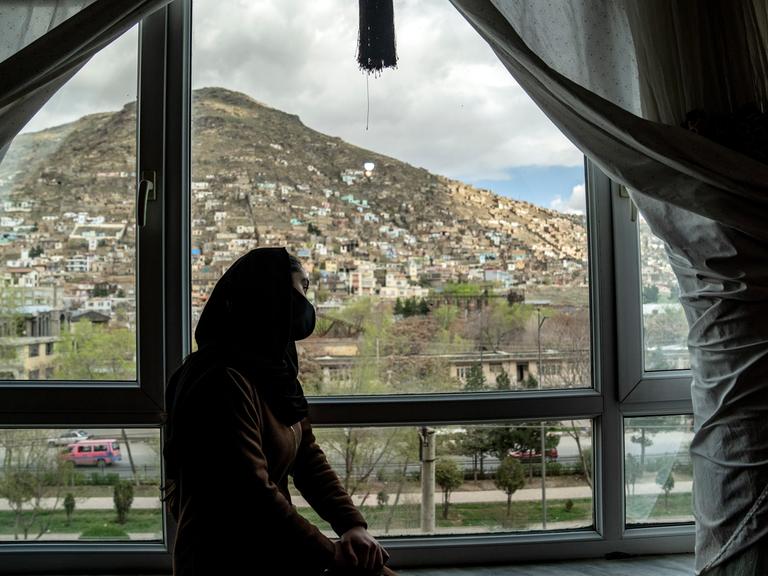 Schattenumriss einer jungen Frau in Afghanistan, die auf einer Fensterbank sitzt und hinausschaut. Im Hintergrund ist eine Stadt zu sehen. 