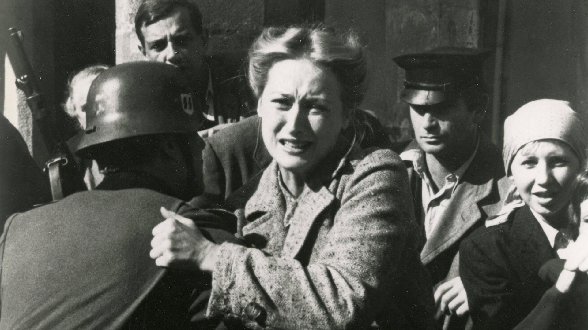 Standfoto aus der Serie "Holocaust", auf dem Meryl Streep von einem Soldaten mit Stahlhelm festgehalten, während sie versucht, sich mit schmerz verzerrtem Gesicht loszureißen.