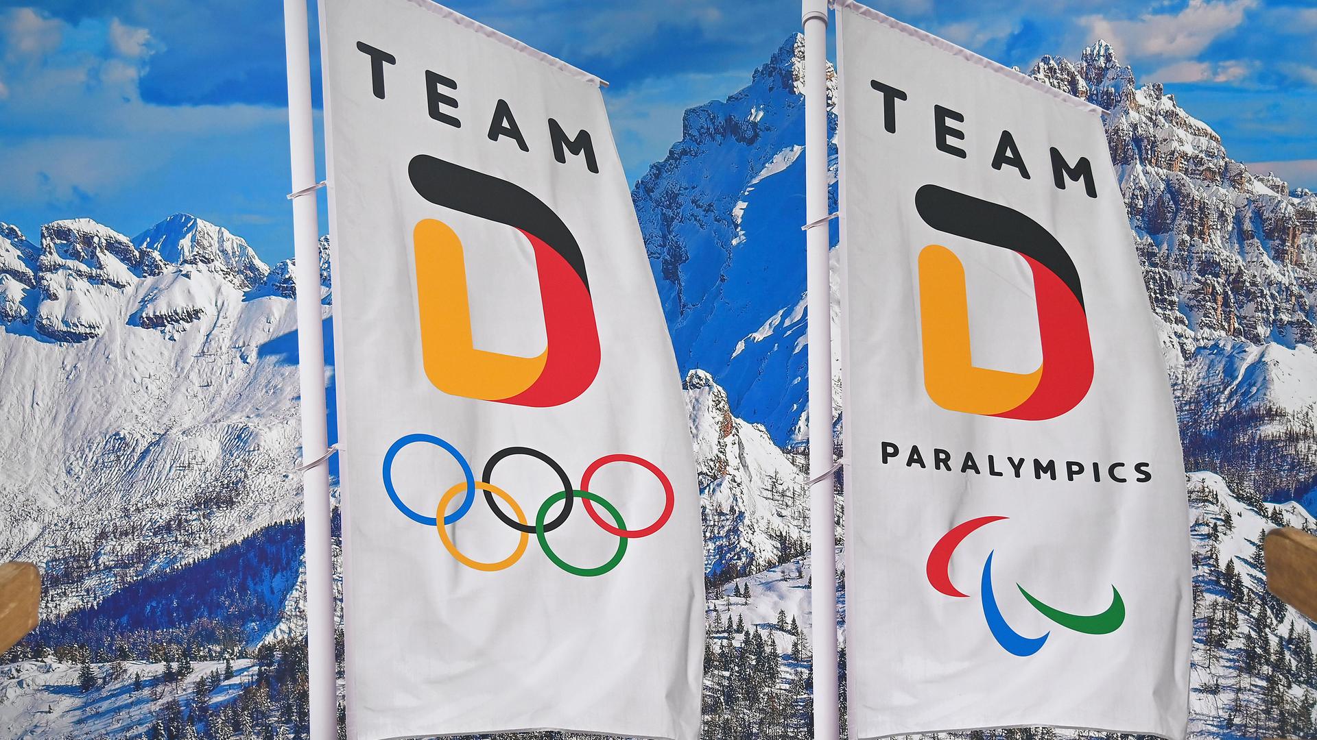 Zwei Fahnen mit der Aufschrift "Team D" wehen vor einer Bergkulisse. Auf der einen Fahne sind die olympischen Ringe abgebildet, auf der anderen Fahne das Logo der Paralympischen Spiele.