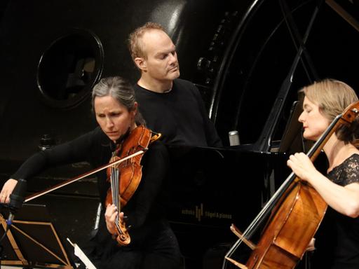Eine Bratschistin, ein Pianist und eine Cellistin musizieren auf einer Bühne. Im Hintergrund sind Maschinen des Jugendstil-Kraftwerks Heimbach (Eifel)  zu sehen.