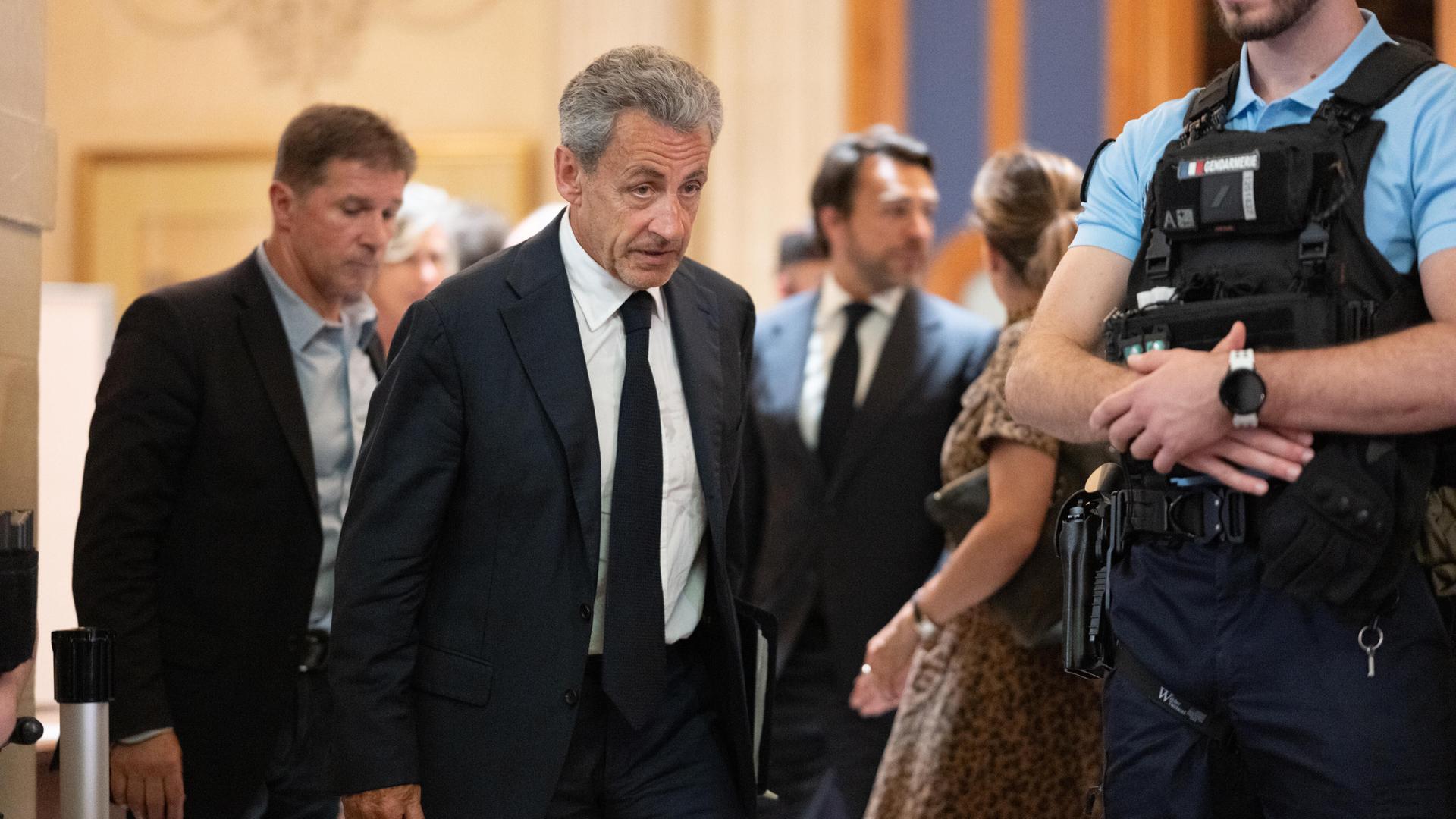 Sarkozy geht im Gerichtssaal an einem Polizisten mit schussicherer Weste vorbei. Hinter ihm weitere Menschen.
