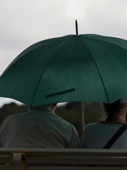 09.08.2024, Hannover, Ein älteres Paar sitzt bei Regen mit grünem Regenschirm auf einer Bank 09.08.2024, Hannover, Ein älteres Paar sitzt bei Regen mit grünem Regenschirm auf einer Bank