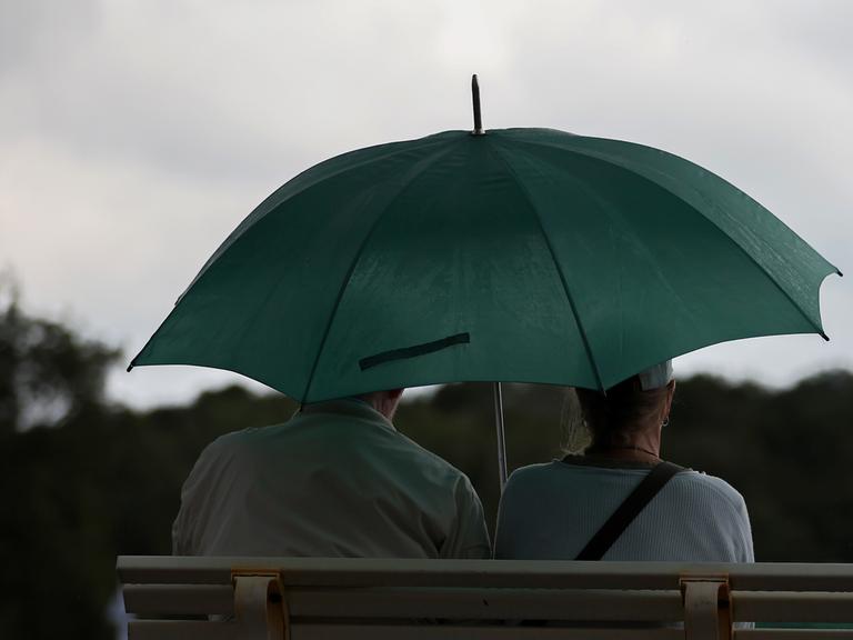 09.08.2024, Hannover, Ein älteres Paar sitzt bei Regen mit grünem Regenschirm auf einer Bank 09.08.2024, Hannover, Ein älteres Paar sitzt bei Regen mit grünem Regenschirm auf einer Bank