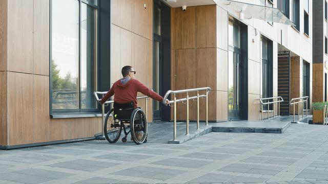 Eine Person in einem roten Kapuzen-Pulli steuert einen Roll-Stuhl über eine glatte Rampe vor einem modernen Gebäude. 