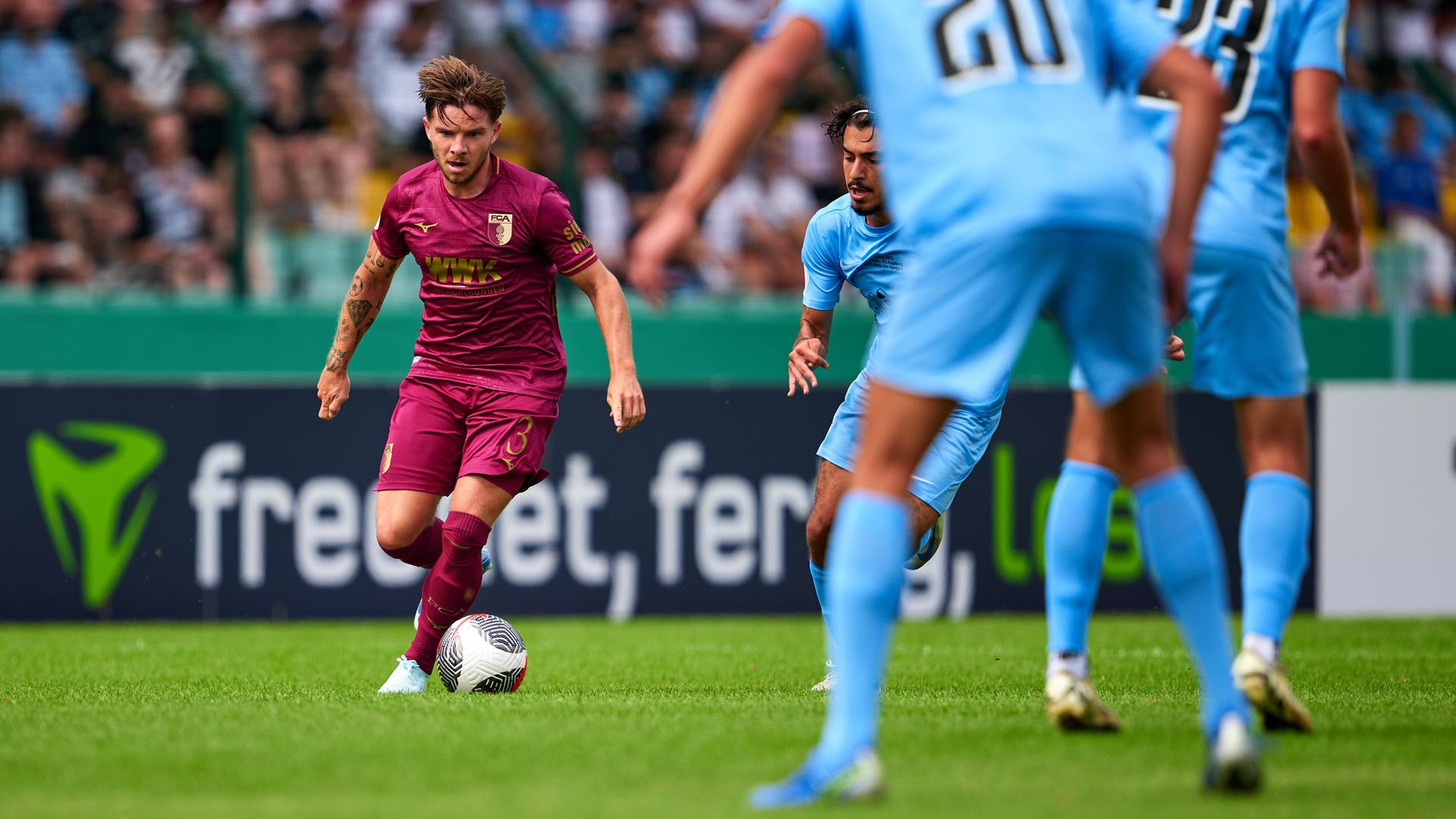 Mads Peterson im roten Trikot auf dem Rasen; im Vordergrund sind mehrere Spieler von Berlin von hinten unscharf zu sehen.