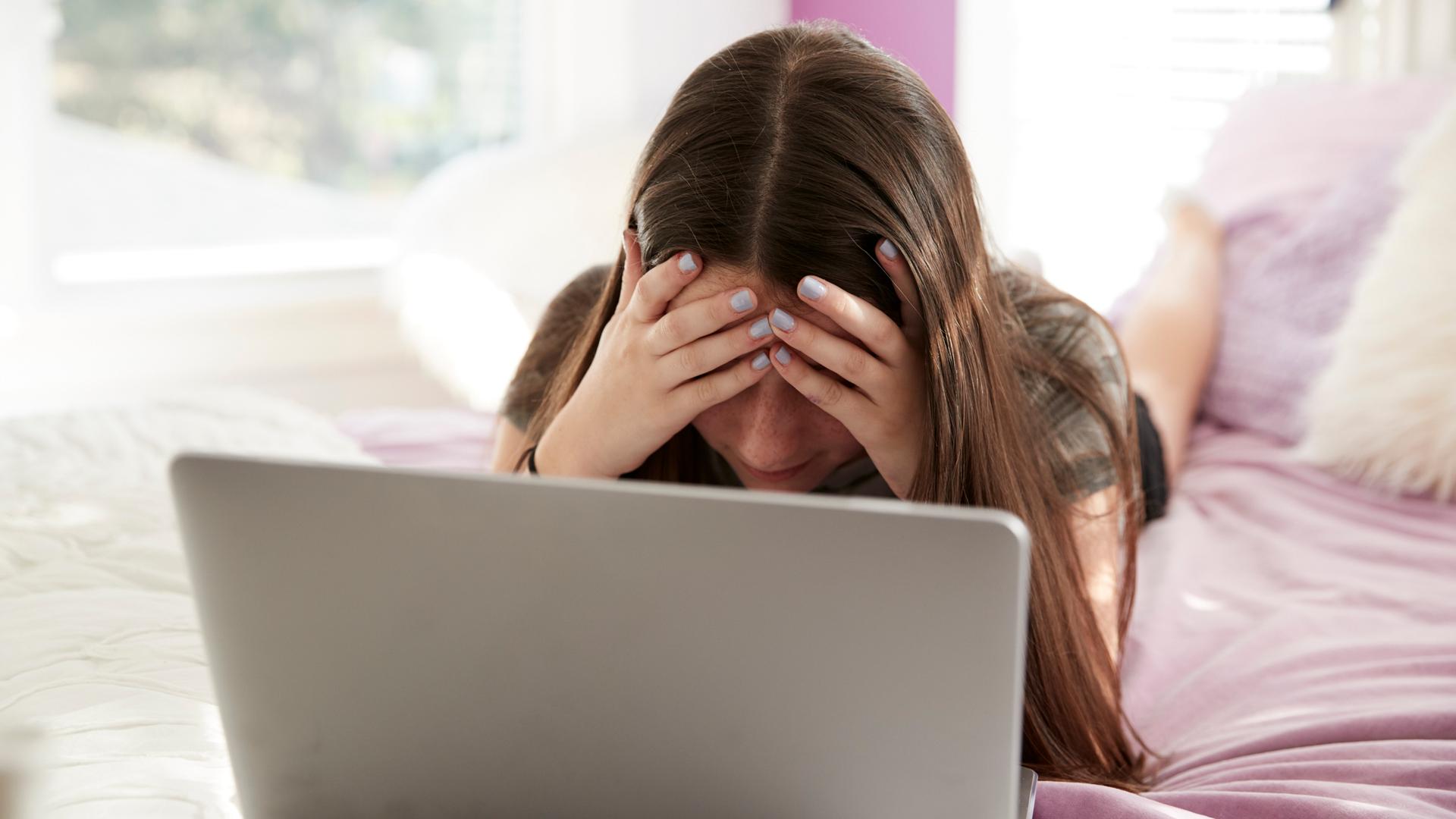 Ein Mädchen mit einem Laptop liegt auf einem Bett. Sie hält sich die Hände vors Gesicht. 