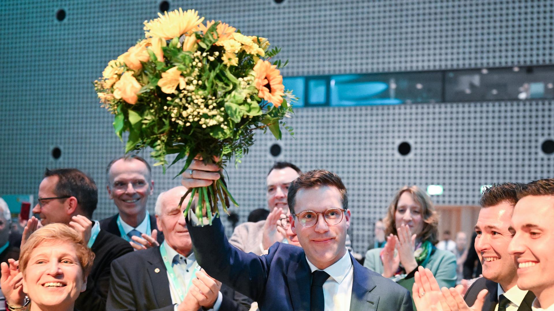 Das Bild zeigt den CDU-Landesvorsitzenden Manuel Hagel auf dem Landesparteitag in Baden-Württemberg. Er hält einen Blumenstrauß in die Höhe, während seine Partei-Kolleginnen und -kollegen ihm zujubeln und Beifall klatschen.