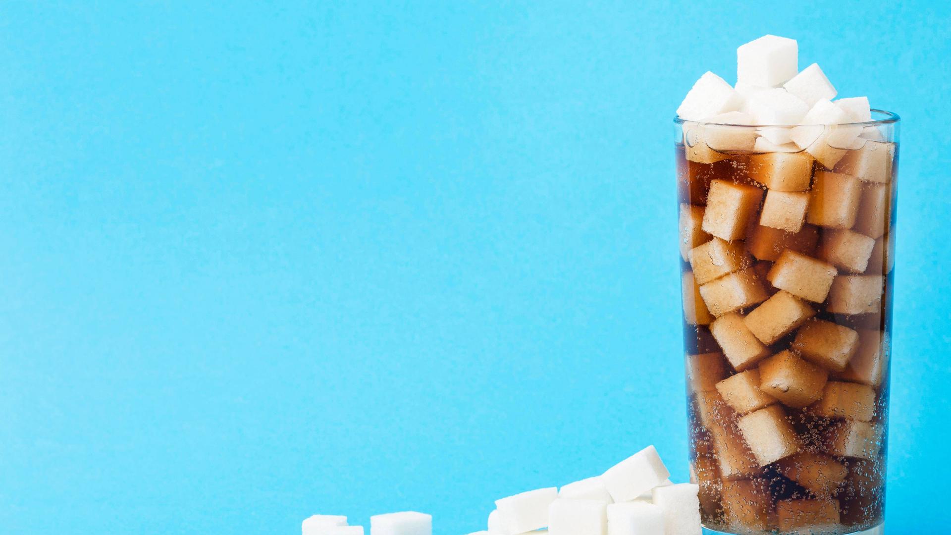 Das stilisierte Bild zeigt ein Glas mit einem Getränk, dem dutzende Zuckerwürfel zugesetzt sind.
