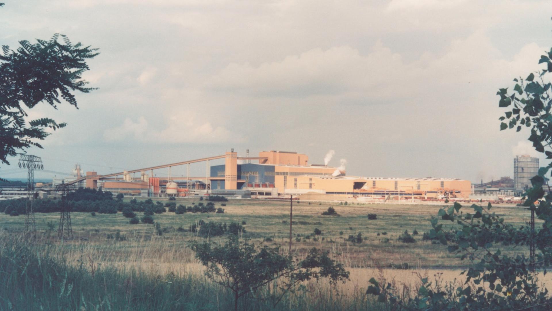 Ein verblichenes Foto vom Eisenhüttenkombinat Ost in Eisenhüttenstadt. Das große Stahlwerk steht an einer Wiese. 