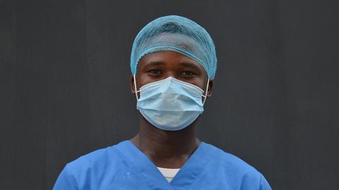 Ein Krankenpfleger in blauer Kleidung, mit Haarnetz und Maske steht vor einer dunkel-grauen Wand.
