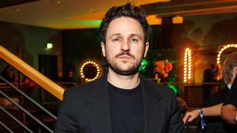 Dimitrij Schaad steht bei einer Veranstaltung im schwarzen Anzug am Fuß einer Treppe und schaut ernst in die Kamera.