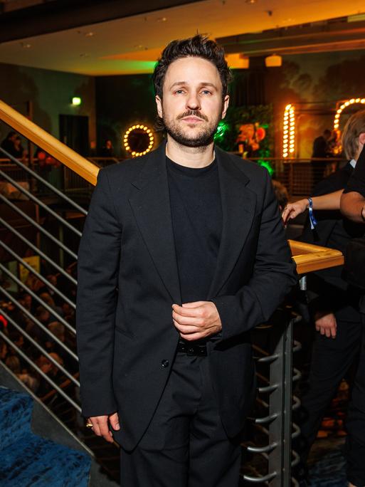 Dimitrij Schaad steht bei einer Veranstaltung im schwarzen Anzug am Fuß einer Treppe und schaut ernst in die Kamera.