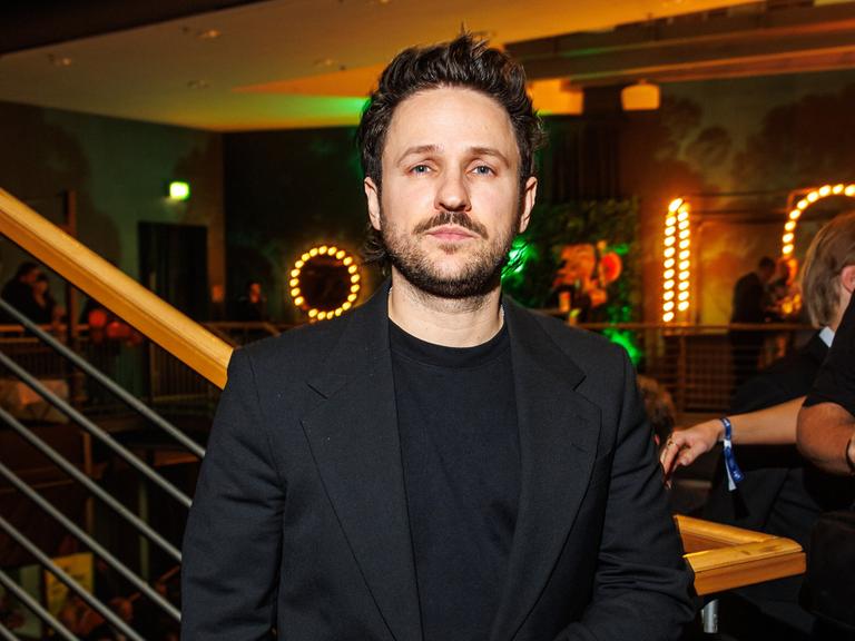 Dimitrij Schaad steht bei einer Veranstaltung im schwarzen Anzug am Fuß einer Treppe und schaut ernst in die Kamera.