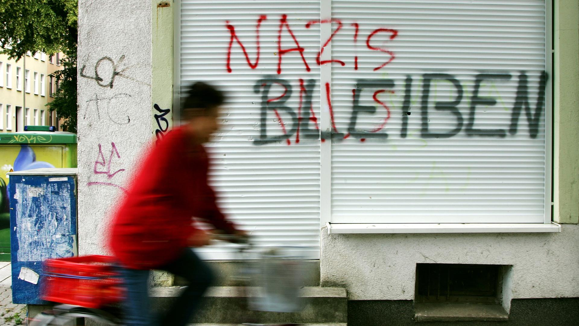 Eine Geschäftsfront ist mit dem roten Schriftzug "NAZIS RAUS", besprüht. Darüber hat jemand in Schwarz "BLEIBEN" geschrieben.