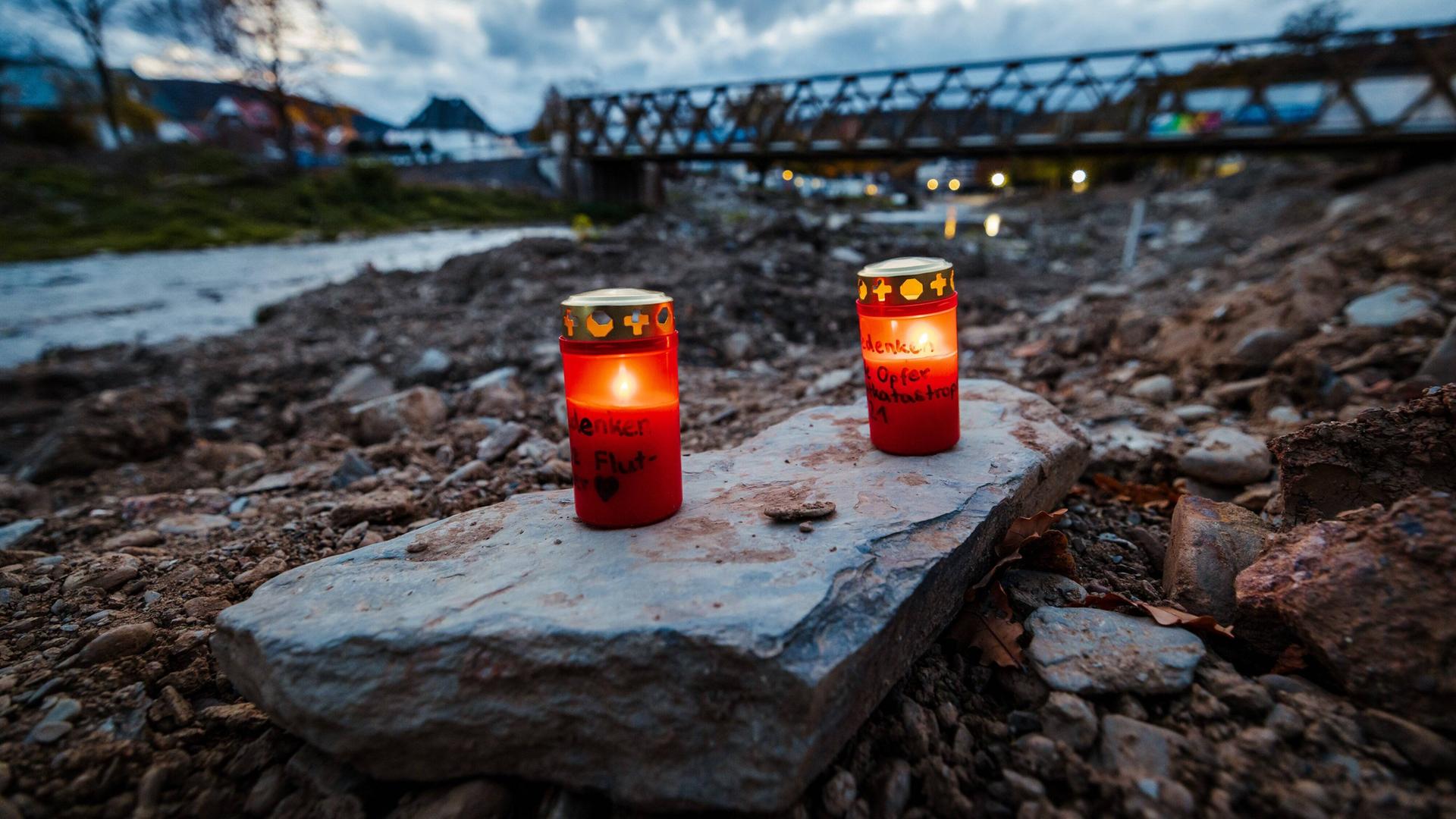 Zwei rote Kerzen stehen auf einem Stein. Im Hinter-Grund sieht man den Fluss und eine Brücke.