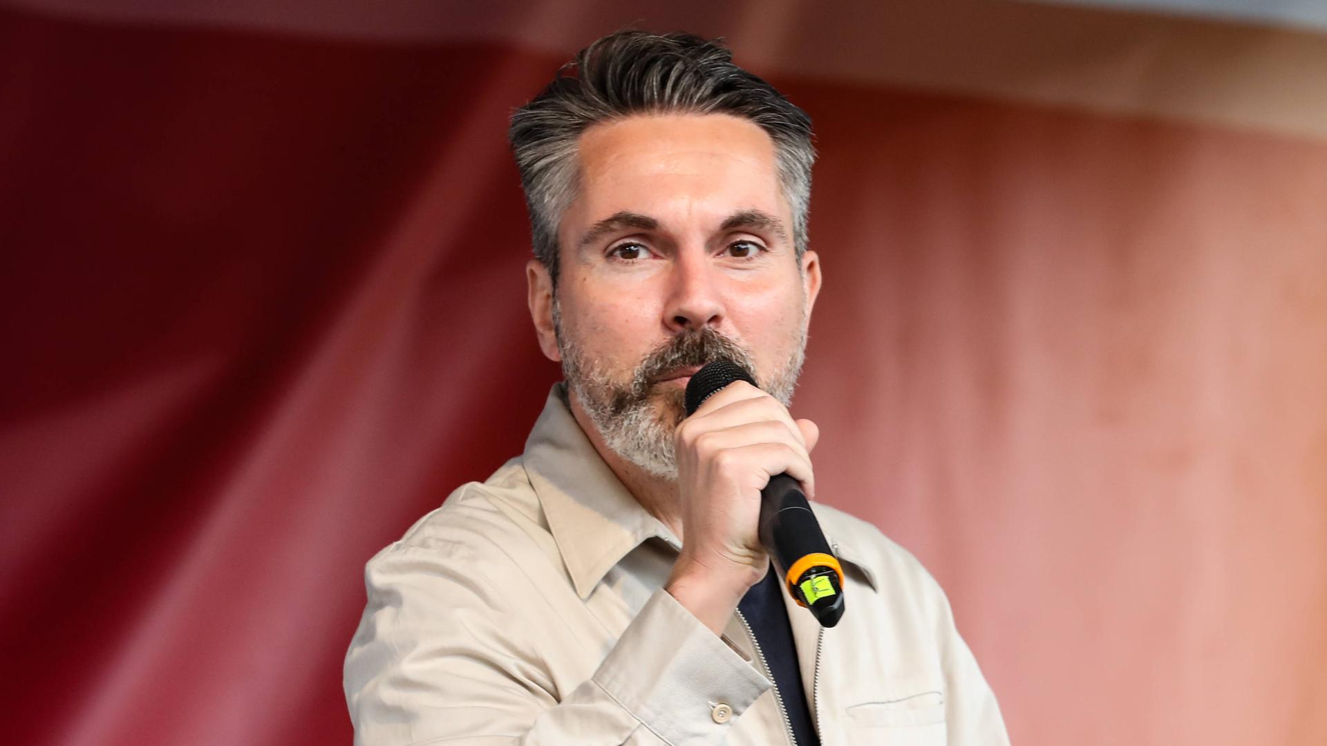 Fabio De Masi spricht in ein Handmikrofon. Im Hintergrund: ein rotes Transparent Fabio De Masi spricht in ein Handmikrofon. Im Hintergrund: ein rotes Transparent