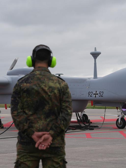 Eine Heron-Drohne der Bundeswehr steht auf einem Flugfeld. Davor ein Soldat mit Gehörschutz.