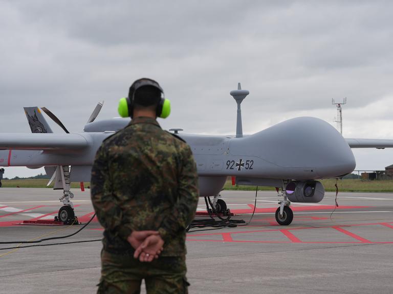 Eine Heron-Drohne der Bundeswehr steht auf einem Flugfeld. Davor ein Soldat mit Gehörschutz.