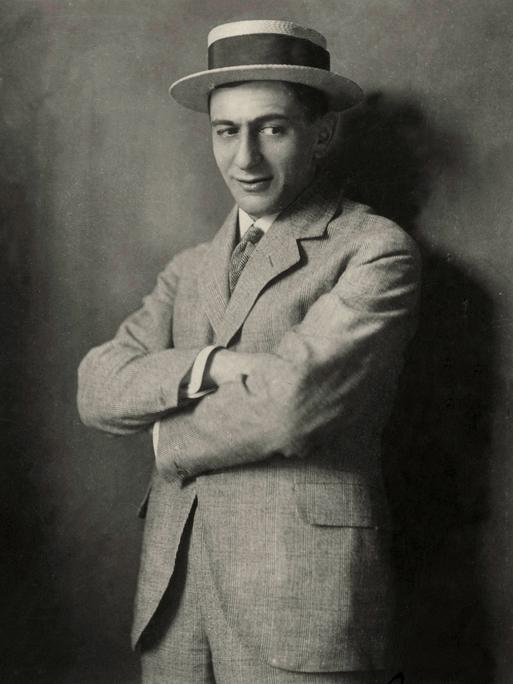 Studioaufnahme von Ernst Lubitsch, der mit verschränkten Armen, leicht lächelnd an der Kamera vorbei schaut. Er trägt Hut.