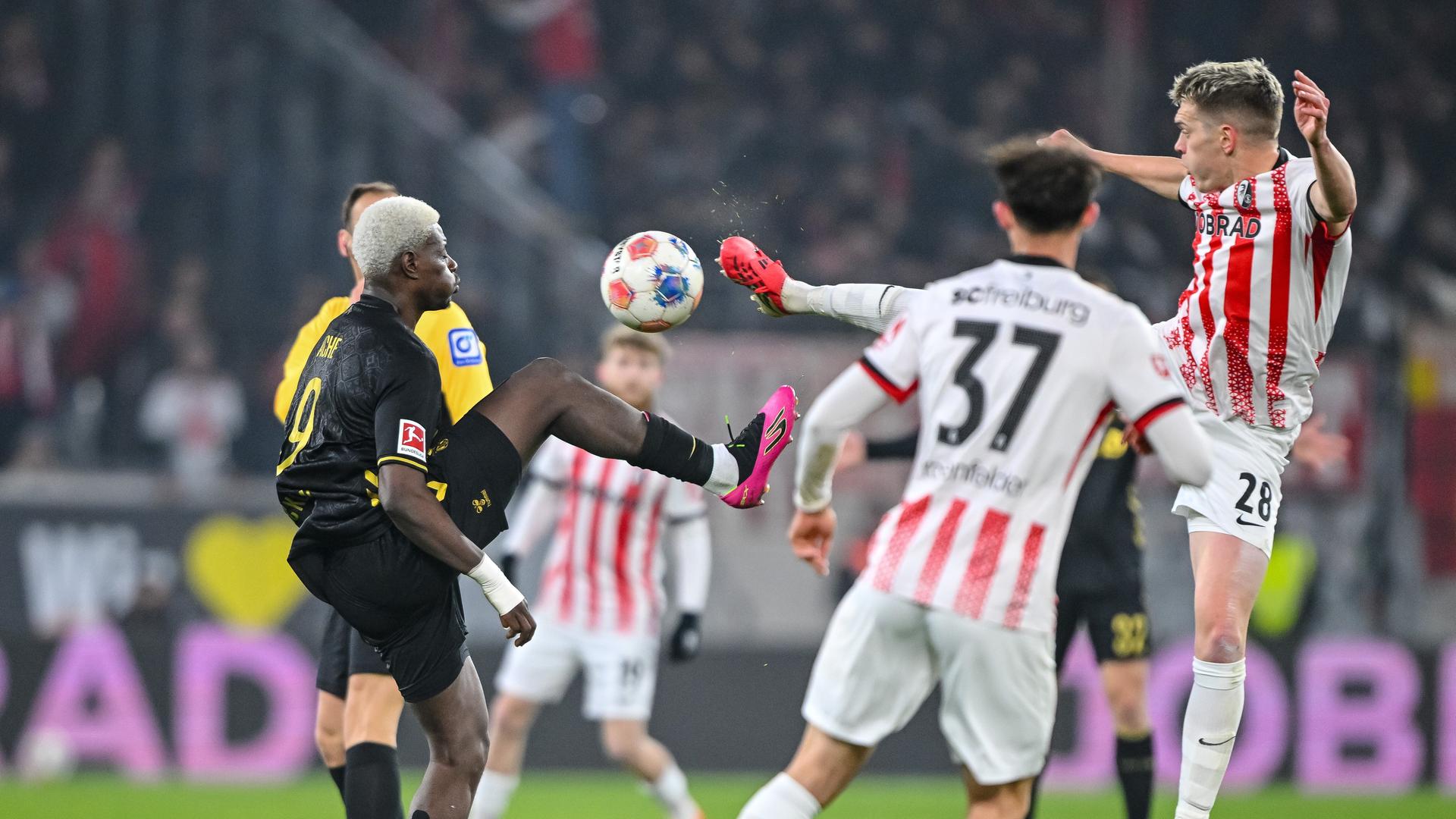 Auf dem Spielfeld kämpfen zwei Spieler mit erhobenen Beinen um den Ball. Das Bild zeigt auf der linken Seite Ragnar Ache im schwarzen Trikot des 1. FC Köln und auf der rechten Seite Matthias Ginter im rot-weiß gestreiften Trikot des SC Freiburg.