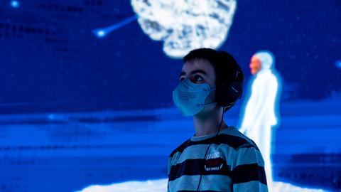 Ein Junge steht in einem Raum, der blau erleuchtet ist, im Hintergrund ist eine Figur zu sehen.