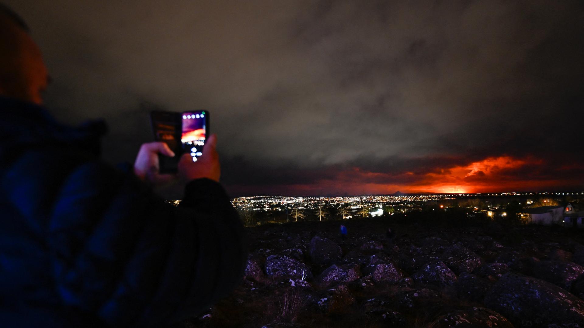 In einem Vorort von Reykjavik machen Menschen mit dem Handy Fotos von dem Vulkanausbruch, der am Horizont zu sehen ist.