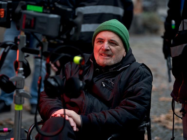 Porträt von Regisseur Uli Edel an einem Filmset. Er sitzt auf einem Stuhl und schaut auf einen Bildschirm. Er trägt eine schwarze Daunenjacke, eine grüne Mütze und einen Drei-Tage-Bart.